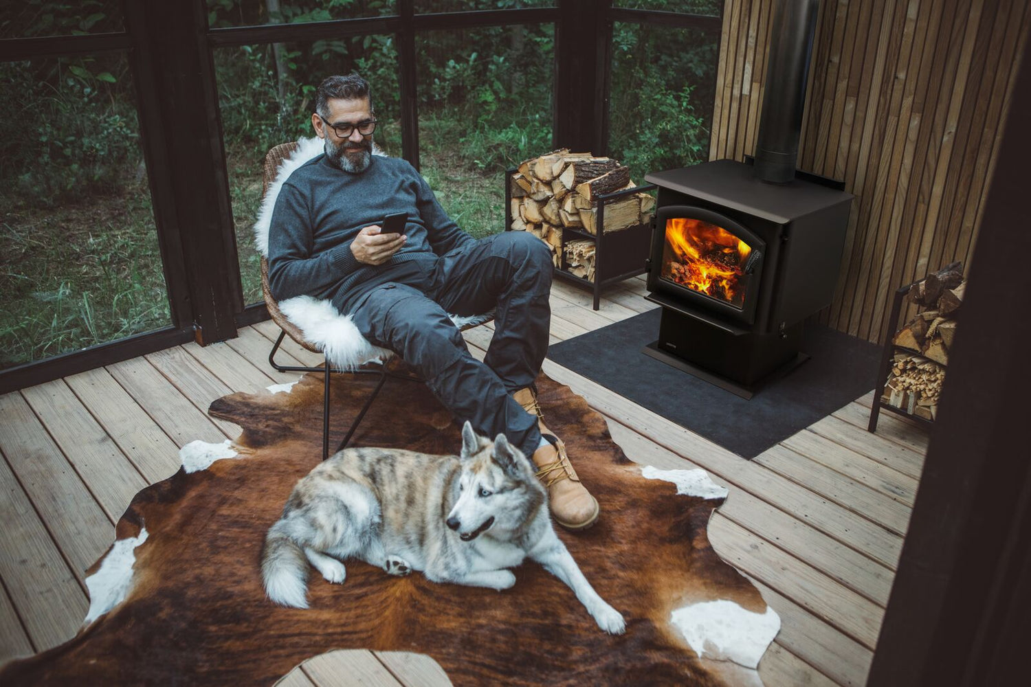 Man seated on a chair, looking at phone, with a dog lying on a fur rug beside a wood stove.
