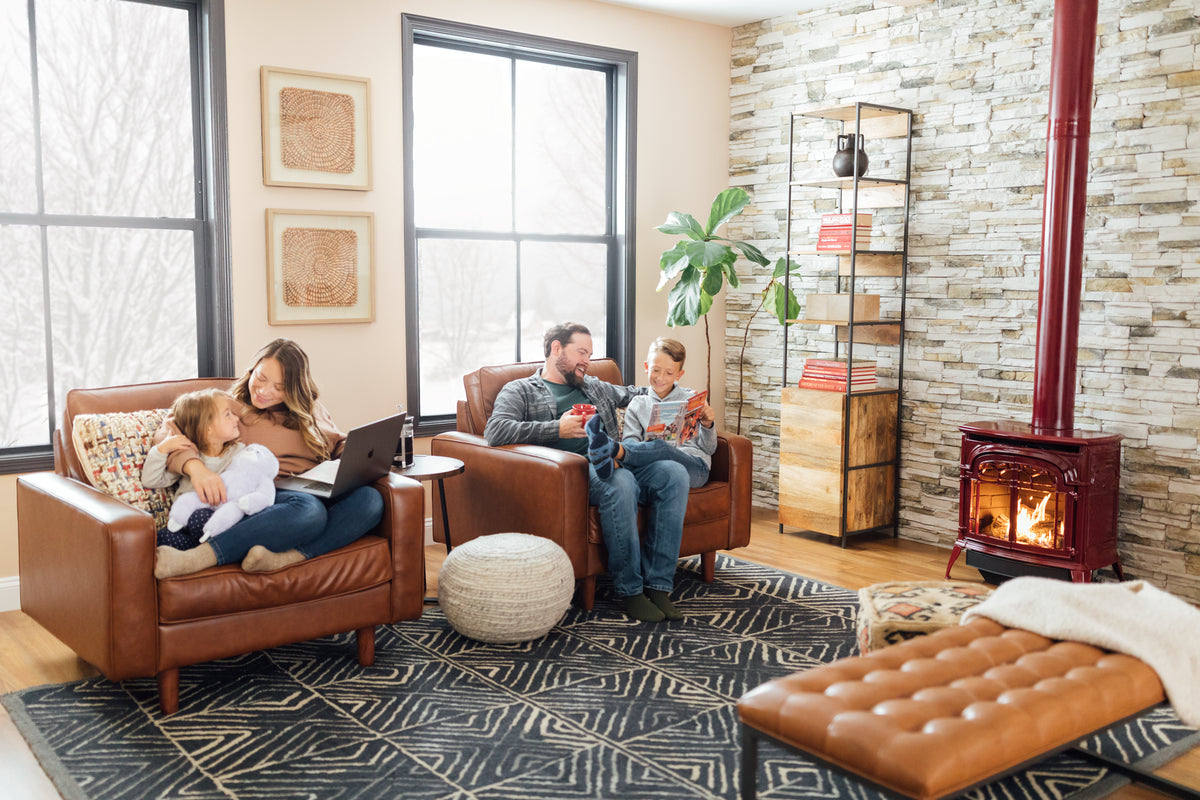 Family enjoys cozy living room; mother with laptop, daughter cuddling; father and son read by wood stove.