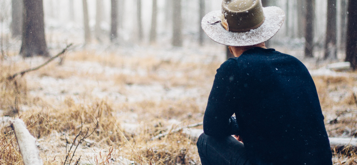 A person wearing a hat sits in a snowy, forested area with tall trees and dry grass.