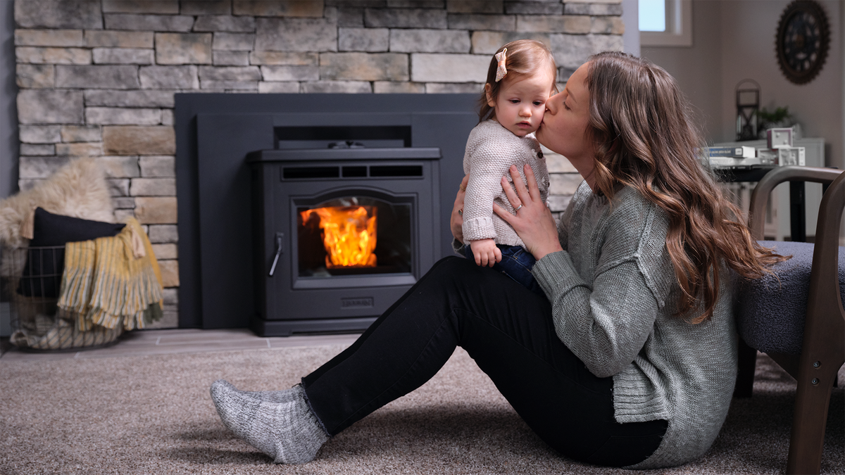 A woman kisses a child beside a lit fireplace insert, in a cozy living room with a stone wall backdrop.